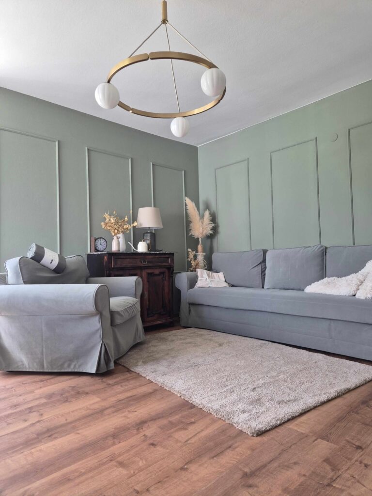 Eingerichteter Wohnraum mit grüner Wandfarbe, Zierleisten, grauem Sofa und Holzfußboden